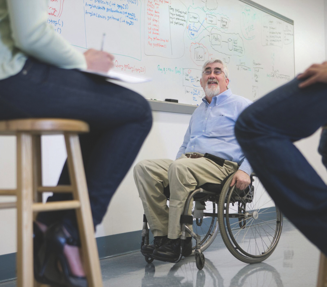 A person in a wheelchair smiling and engaging in a discussion with others in a classroom with whiteboards covered in notes and diagrams.